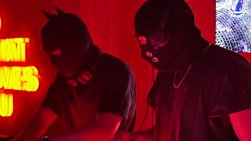 Under red lighting, two DJs wearing masks and headphones play bass music in Cebu, with a disco ball and a glowing sign visible. - midnightrebels.com