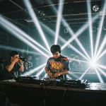 A DJ performs under bright lights at a nightclub, surrounded by photographers capturing the event. The scene illustrates the symbiotic relationship between nightlife and photography, where photographers document the energy and atmosphere of the music scene. - midnightrebels.com