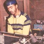 Vintage photo shows a DJ with a blue headband and striped shirt operating turntables and a mixer, with vinyl records stacked nearby. - midnightrebels.com