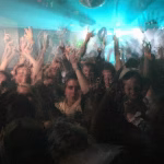 A blurry, low-lit photograph captures a crowded rave scene with people raising their hands under a disco ball and bright blue lights. - midnightrebels.com