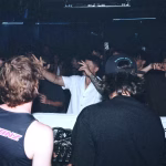 A DJ stands behind a mixer at a crowded, dimly lit nightclub, with partygoers visible in the background. - midnightrebels.com