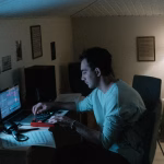 A musician composes music on a laptop using Ableton Live or FL Studio, utilizing a midi controller in a dimly lit bedroom studio. - midnightrebels.com