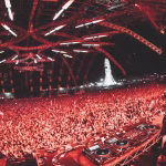 A wide shot captures a DJ onstage with their equipment overlooking a massive crowd lit in red, under a complex lighting rig, with the Coachella Ferris wheel visible in the background, symbolizing the festival's large scale and electronic music presence. - midnightrebels.com