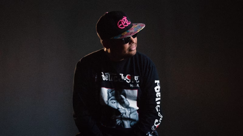 JaCas, the nightlife photographer, is pictured wearing sunglasses, a patterned-brim cap, and a black long-sleeved shirt with a Tupac Shakur graphic, against a dark background. - midnightrebels.com