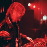 A female DJ in a leather jacket, illuminated by red lighting, performs at a club console with a crowd visible in the background. - midnightrebels.com
