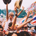 A crowd of cheering festival-goers, some with hands raised and a woman on someone's shoulders, celebrate with a Thai flag and a flag representing multiple countries visible. - midnightrebels.com
