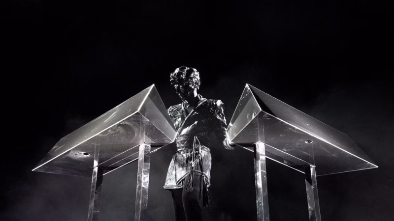 Gesaffelstein, shrouded in mystery, performs on stage behind two pyramidal metallic consoles against a stark black backdrop, illustrating his enigmatic presence in the techno scene. - midnightrebels.com