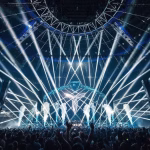 A large crowd enjoys a concert with bright white lasers and stage lighting illuminating a stage with the AXSOM logo. - midnightrebels.com