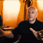 DVS1, a bald man in a black shirt, gestures with his hands in his music studio, surrounded by synthesizers and audio equipment, in a warm-toned, candid shot for an article about his views on hard techno. - midnightrebels.com