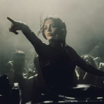 Female DJ pointing in crowded, smoky club with Pioneer decks.
