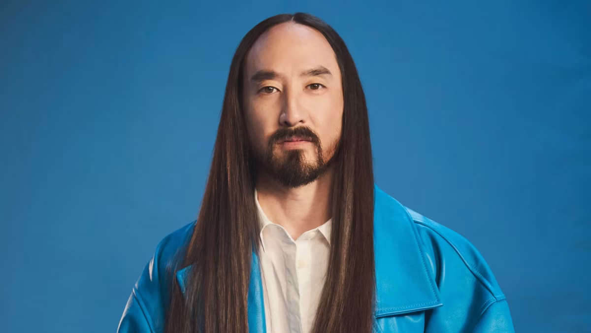 The image shows a portrait of DJ Steve Aoki with long dark hair, a beard, and a blue jacket, against a blue background. - midnightrebels.com