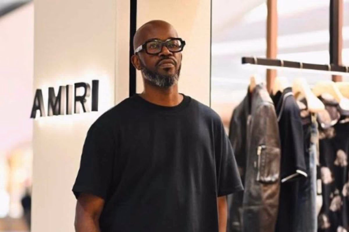 DJ Black Coffee, wearing a black t-shirt and glasses, stands in front of an Amiri store display. - midnightrebels.com