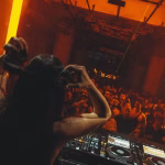 DJ performing for crowd in nightclub, warm orange lighting.