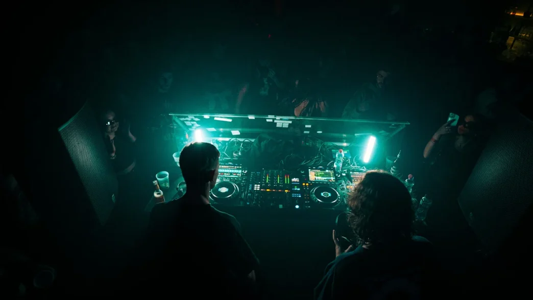Two DJs performing a live set at a dark, atmospheric nightclub with teal neon lighting, using a Pioneer DJ controller and professional sound system in front of a dancing crowd.