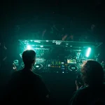 Two DJs performing a live set at a dark, atmospheric nightclub with teal neon lighting, using a Pioneer DJ controller and professional sound system in front of a dancing crowd.