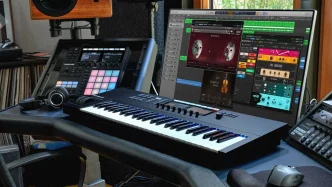 A music production studio featuring Native Instruments hardware, including a Komplete Kontrol keyboard and Maschine controller, alongside a monitor displaying software like Guitar Rig.