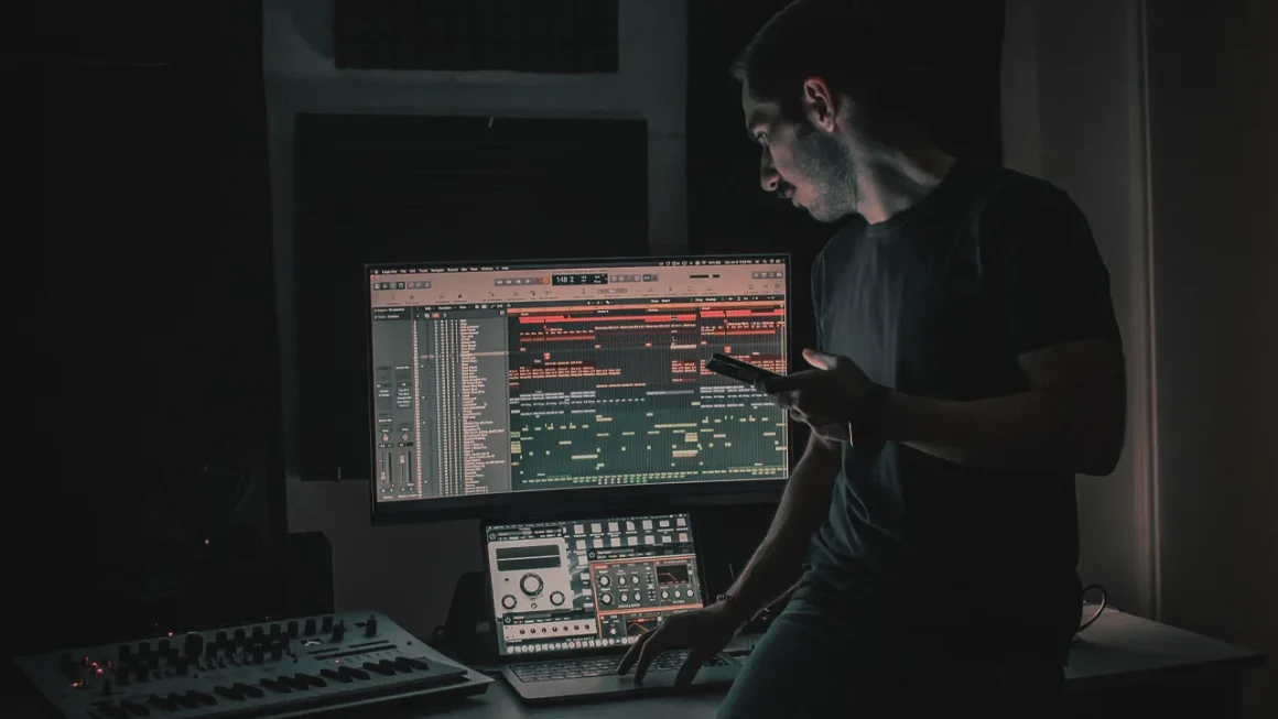 Music producer working at night on laptop with professional synthesizer and audio equipment controlling DAW
