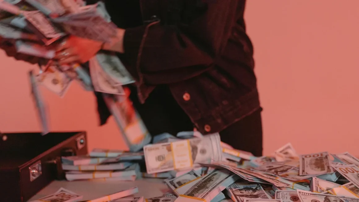 Person in black jacket handling large stacks of US hundred-dollar bills on a table