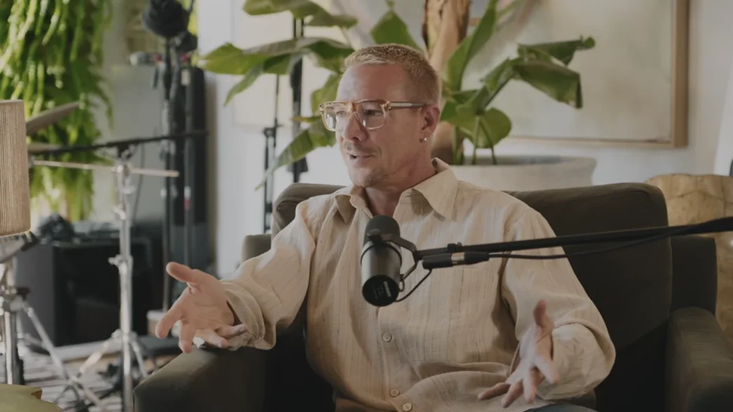 Diplo wearing glasses and a light beige shirt speaking into a microphone during an interview or podcast in a room with plants and musical equipment