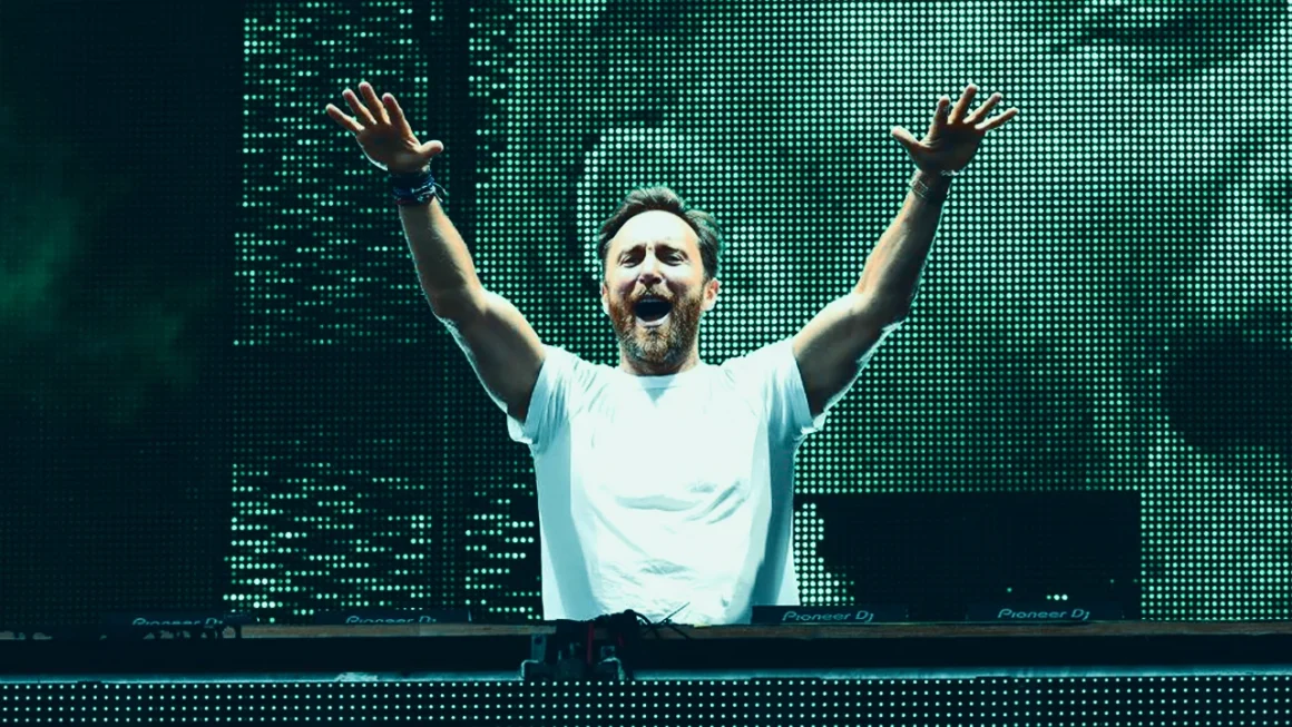 David Guetta performing with arms raised on stage in front of LED screen