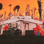 Performers in red outfits dancing on a stage with a building and communication antennas under an orange sky alongside Major Lazer at Coachella 2026