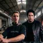Two men standing in an industrial warehouse with DJ equipment, one adjusting controls and the other looking forward