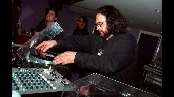A DJ manipulating vinyl records and a mixer at a lively event, with attendees in the background.