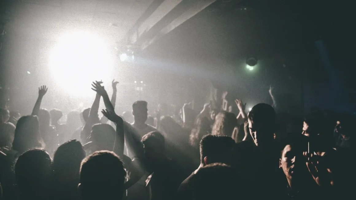 People dancing and raising hands in a dimly lit crowded club with bright stage lights