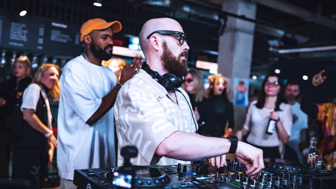DJ wearing headphones and adjusting controls on a mixer at a lively party with people dancing and socializing in the background