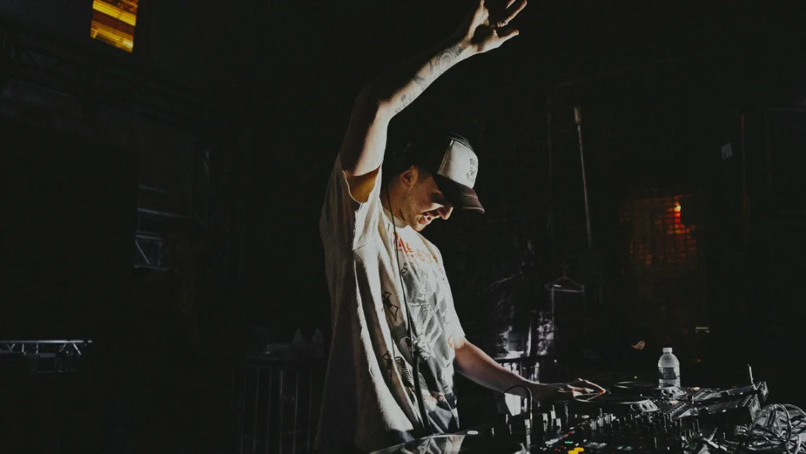 DJ wearing a cap and headphones, adjusting controls on a mixing console with one hand raised