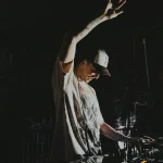 DJ wearing a cap and headphones, adjusting controls on a mixing console with one hand raised