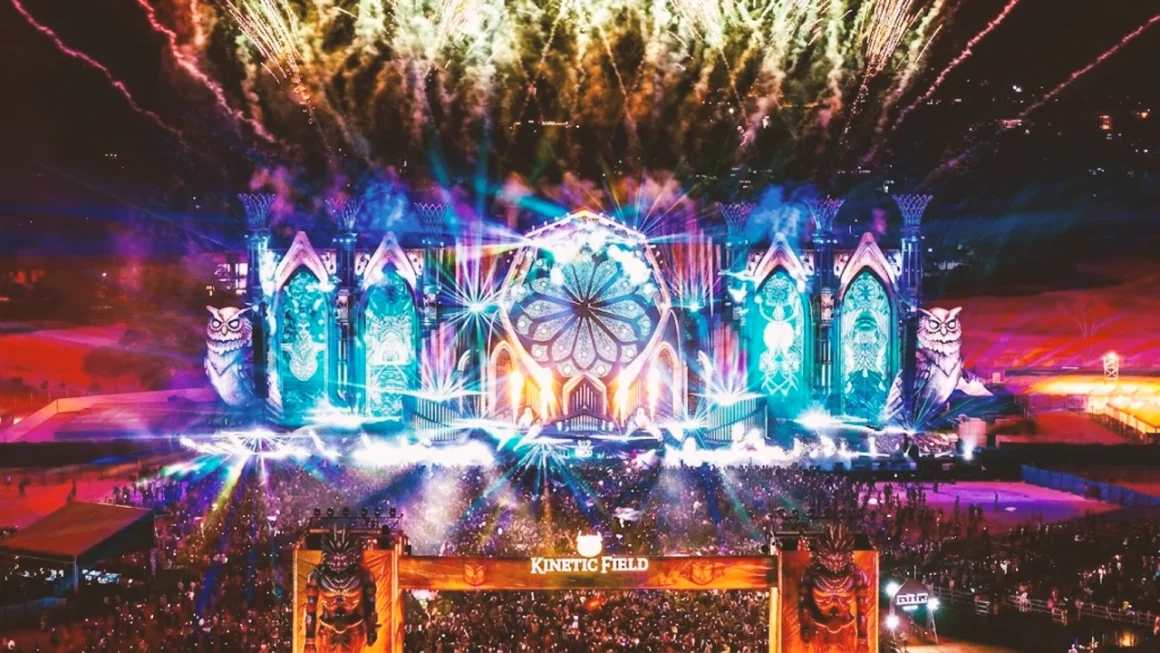 Fireworks explode over Kinetic Field festival stage, owls, and large crowd.
