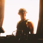 Smiling DJ in visor performing electronic music on a bright stage.