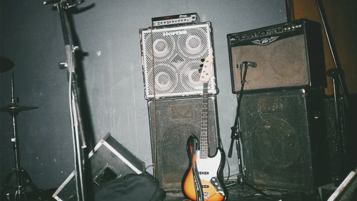 Grungy live music stage with bass guitar and huge amplifier stacks.