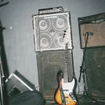 Grungy live music stage with bass guitar and huge amplifier stacks.