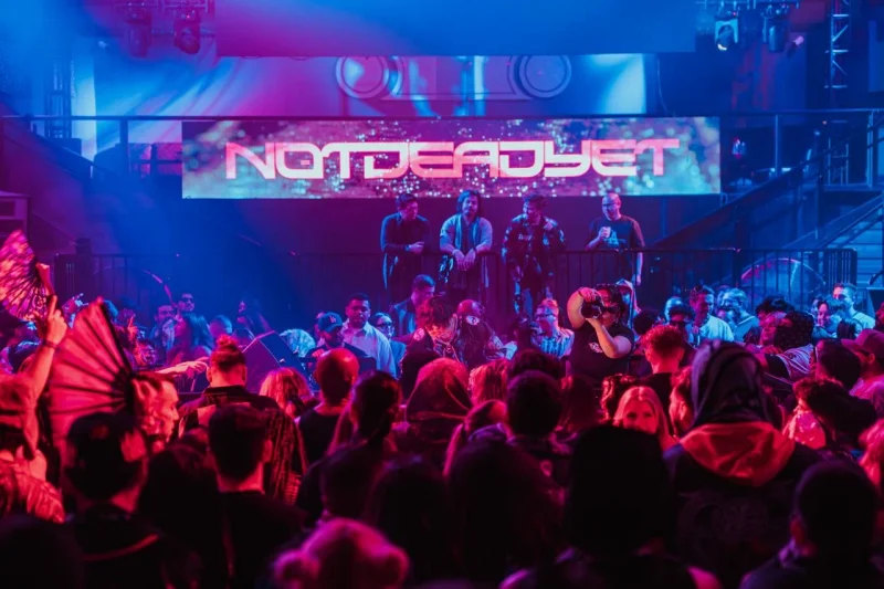 Lively crowd enjoys 'NOTDEADYET' concert under vibrant blue and red lights.