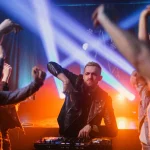 Energetic DJ performs for dancing crowd under vibrant blue and orange club lights.