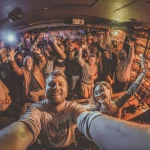 Smiling DJ and happy crowd taking a selfie at an energetic nightclub.