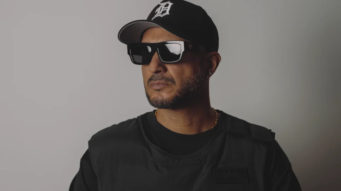 Man in black D-logo cap, sunglasses, and Alpha Industries vest.