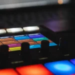 Electronic music drum machine with colorful backlit pads, screen, and controls.