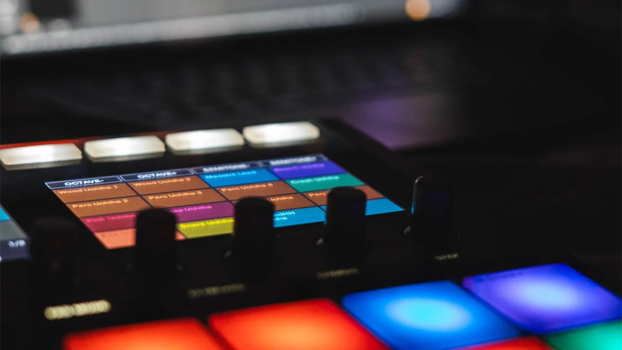 Electronic music drum machine with colorful backlit pads, screen, and controls.