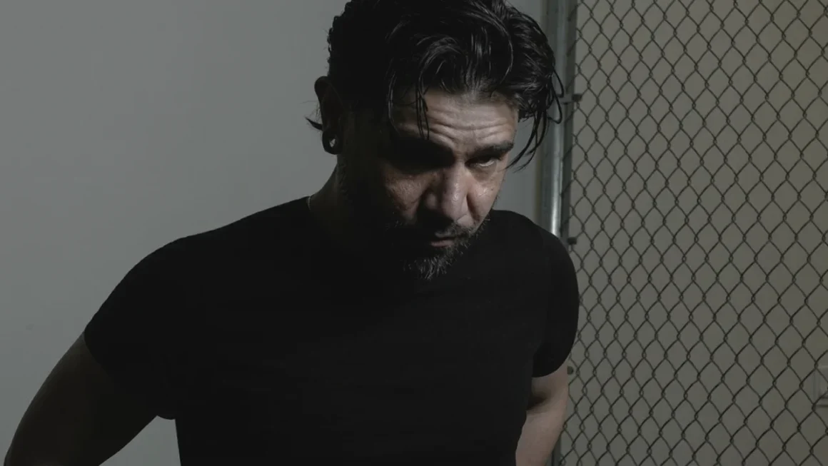 Serious man with beard and black shirt looks down by chain link fence.