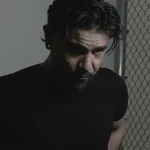 Serious man with beard and black shirt looks down by chain link fence.